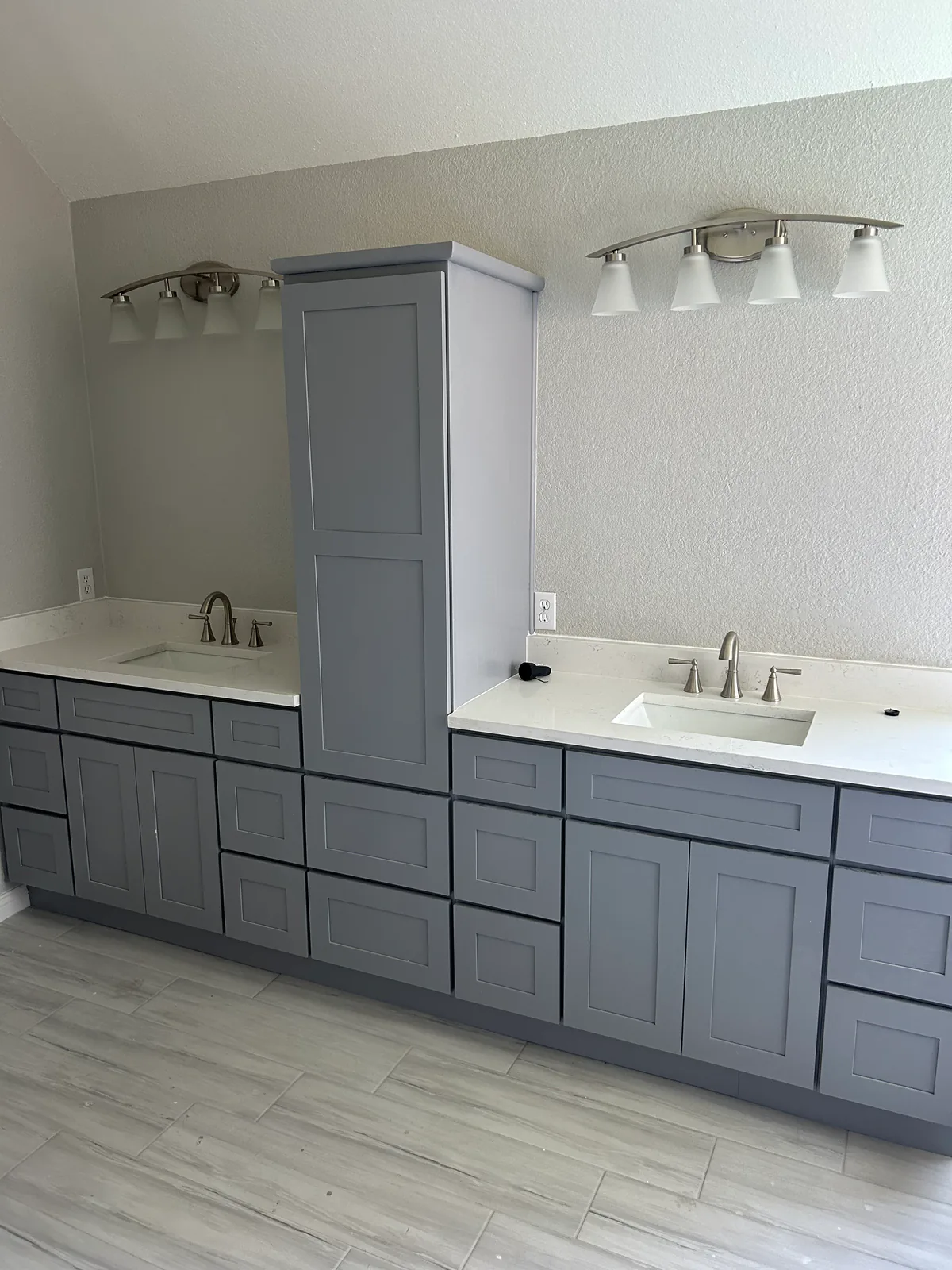 A finished bathroom remodel by AA Flooring and Design — grey shaker double vanity with white quartz countertops, undermount sinks and wood-look plank tile flooring